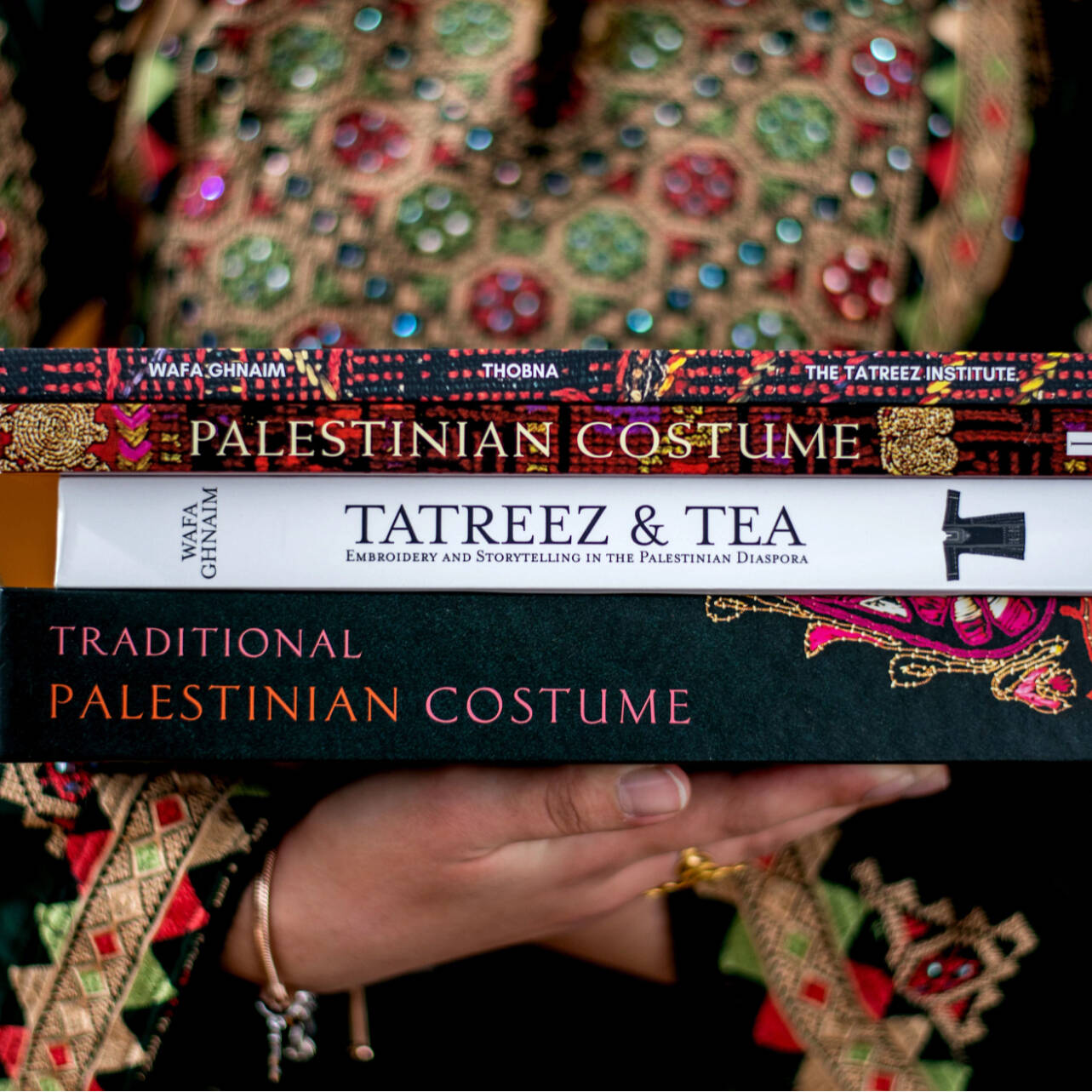 A student wearing traditional Palestinian dress, holding various books, some titles including "Palestinian Costume" and "Traditional Palestinian Costume"